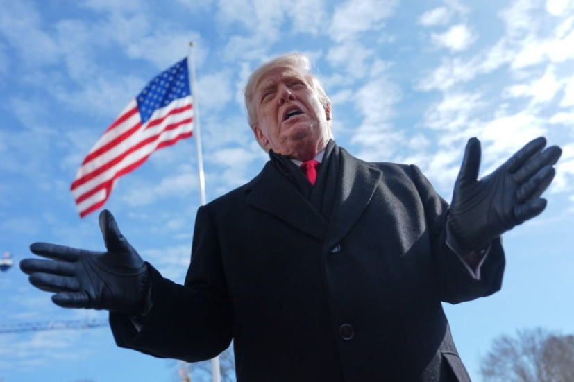 Donald Trump, wearing a black overcoat and gloves with a strong expression, gestures outward with open hands, set against a background of the American flag under a sunny sky. The image captures the decisive tone matching his statement where Trump predicts swift reopening of the Strait of Hormuz.